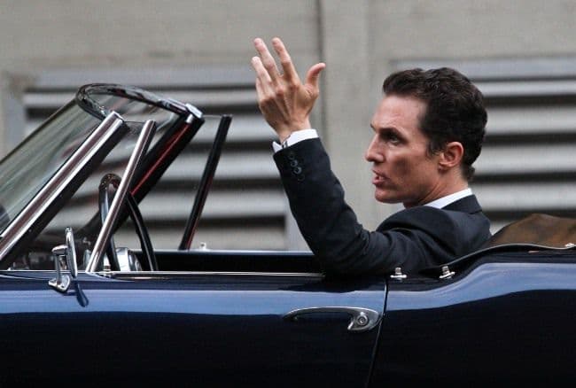 A man in a suit waving from the driver's seat of an open-top car
